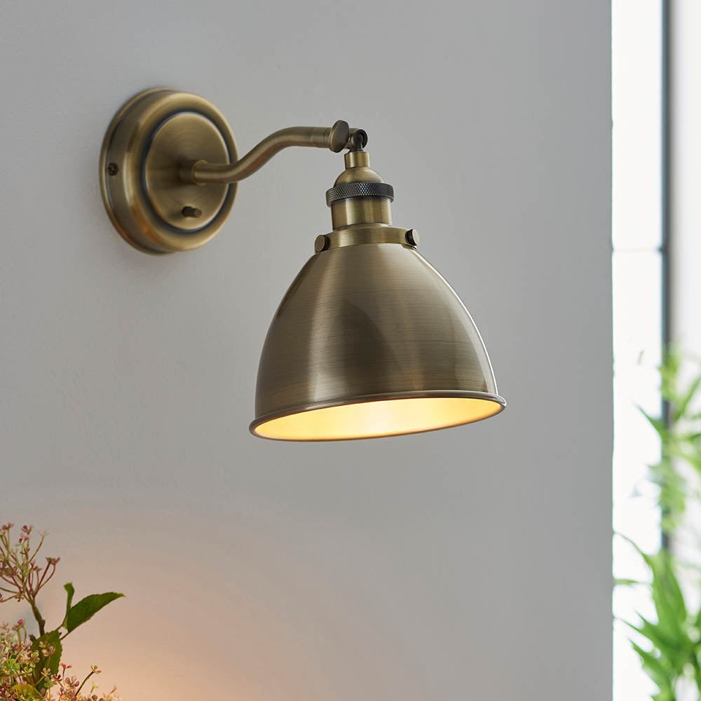 Endon Bramdale industrial wall light in antique brass with adjustable knuckle and domed metal shade, mounted on a pale grey wall.