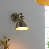 Endon Bramdale wall light in antique brass with adjustable knuckle and domed shade, mounted on a light wall in a bright interior setting.