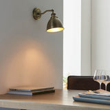 Endon Bramdale wall light in antique brass with adjustable knuckle and dome shade, mounted above a wooden dining table with laptop and wine glasses.