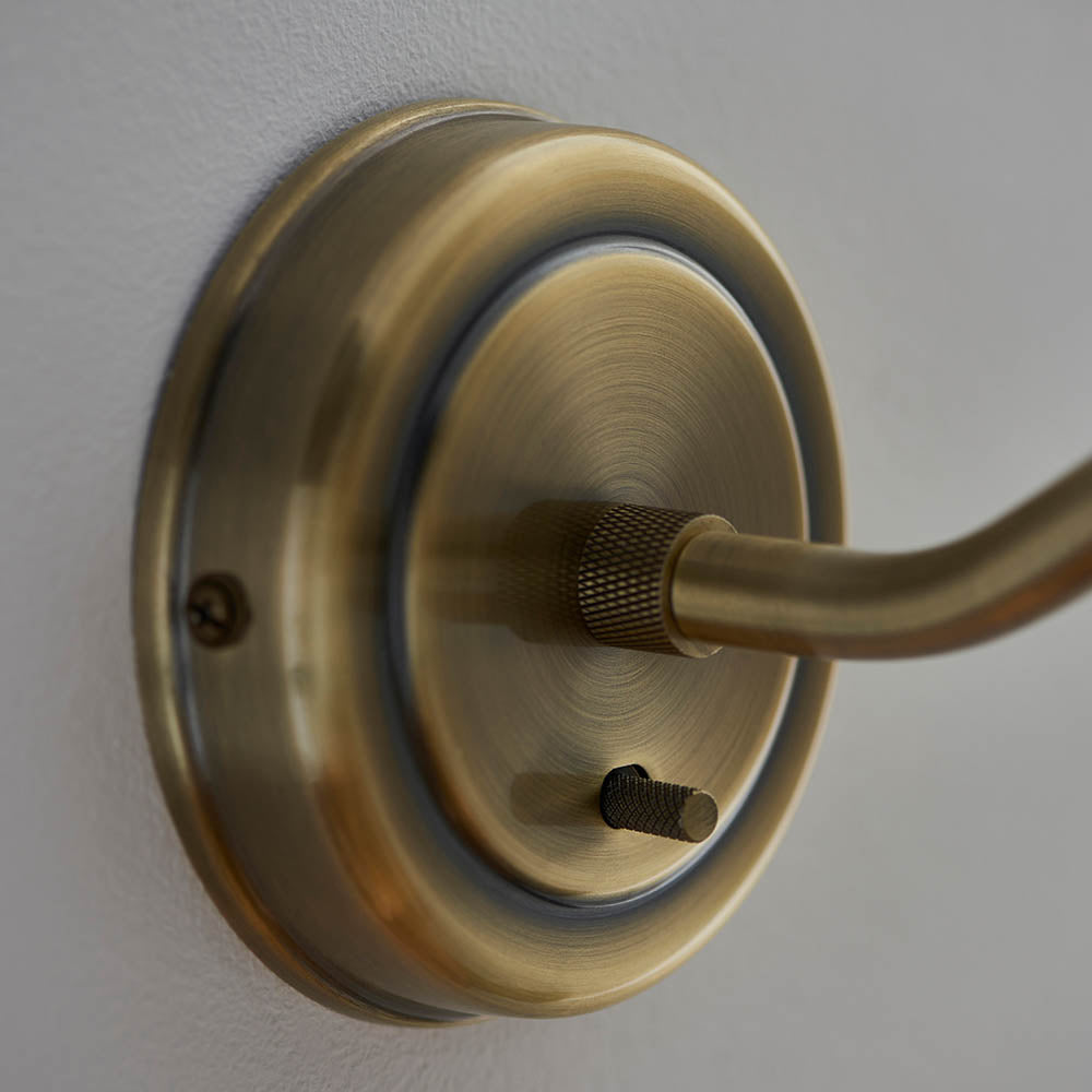 Close-up of Endon Bramdale wall light in antique brass, showing the circular backplate, knurled adjustable knuckle and toggle switch on a pale wall.