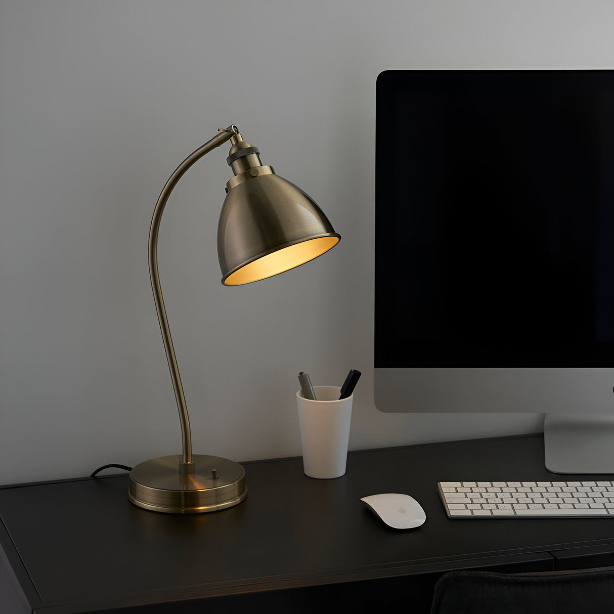Endon Bramdale table lamp in brass with an adjustable domed shade and curved arm, shown on a dark desk beside a monitor, keyboard and pen pot.