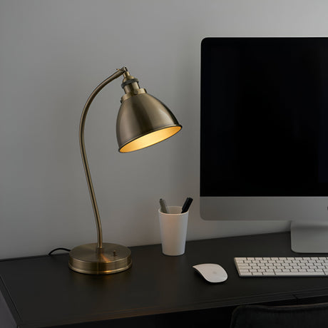 Endon Bramdale table lamp in brass with an adjustable domed shade and curved arm, shown on a dark desk beside a monitor, keyboard and pen pot.