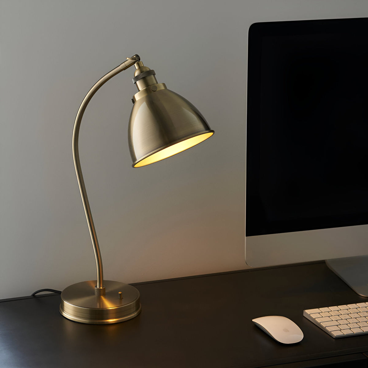 Endon Bramdale brass table lamp with adjustable domed shade and arched stem, lit on a dark desk beside a monitor, keyboard and mouse.
