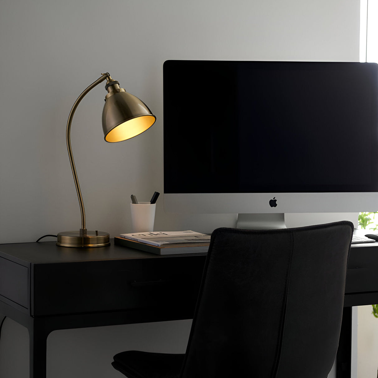 Endon Bramdale brass table lamp with curved arm and dome shade illuminated on a black desk beside an iMac in a modern home office.