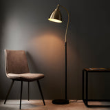 Endon Bramdale floor lamp with an adjustable brass dome shade on a black stem and base, beside a brown chair and side table in a dim room.