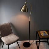 Endon Bramdale industrial floor lamp with antique brass curved arm and domed shade on a matt black stem, beside a grey chair and side table.