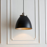 Endon Bramdale medium pendant light with matt black rolled-edge metal shade, aged pewter detailing and inner, on black flex against white panelled wall.