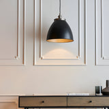 Endon Bramdale medium pendant light with matt black rolled-edge metal shade, aged pewter detailing and inner, hung on black flex against panelled wall.