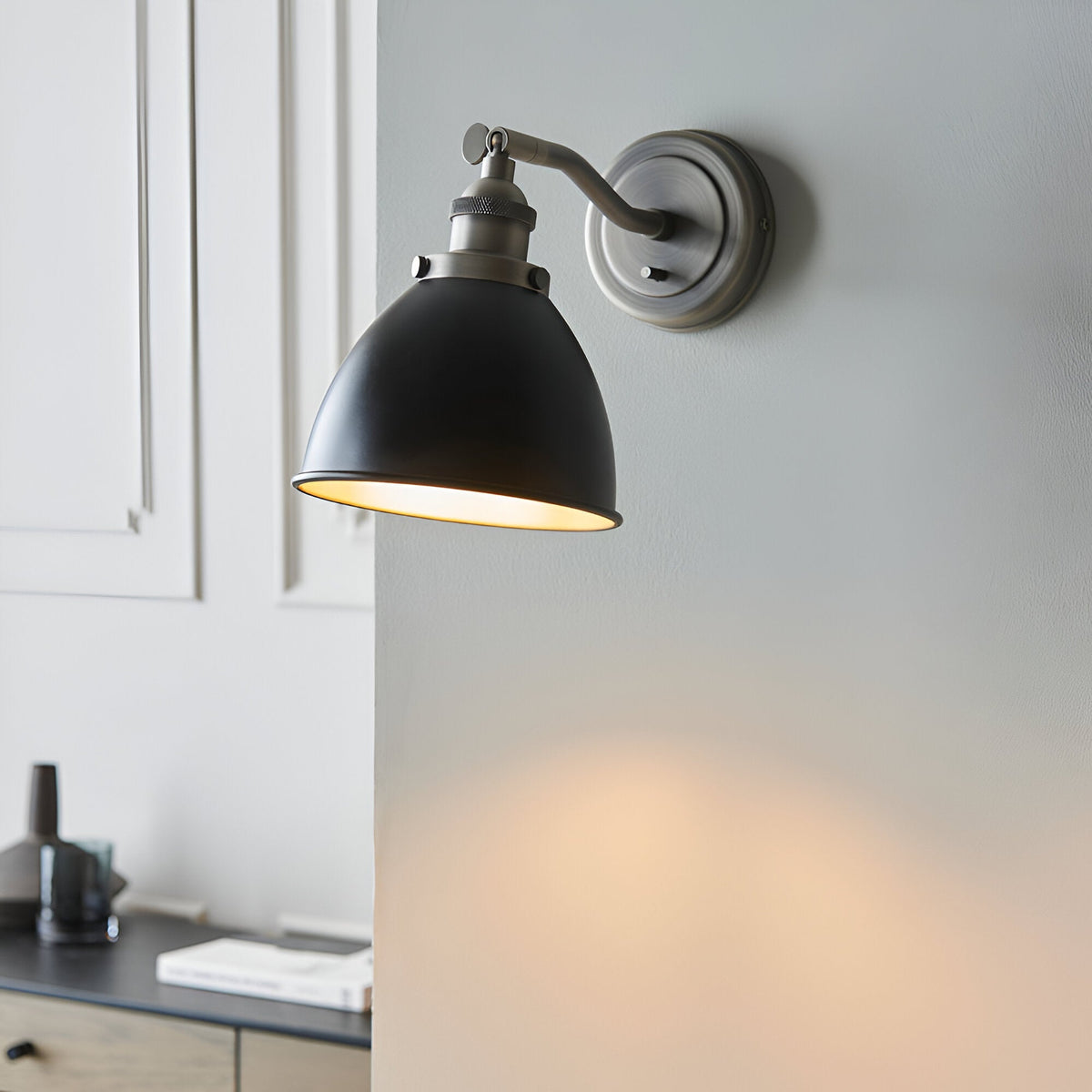 Endon Bramdale industrial wall light with adjustable aged pewter arm and matt black dome shade, mounted on a grey wall beside white panelling.
