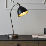Endon Bramdale table lamp with adjustable matt black domed shade and aged pewter curved arm on a wooden sideboard against a panelled grey wall.