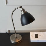 Endon Bramdale table lamp with curved silver metal arm, adjustable knuckle, matt black rolled-edge shade and toggle switch, on a dark desk beside a book.