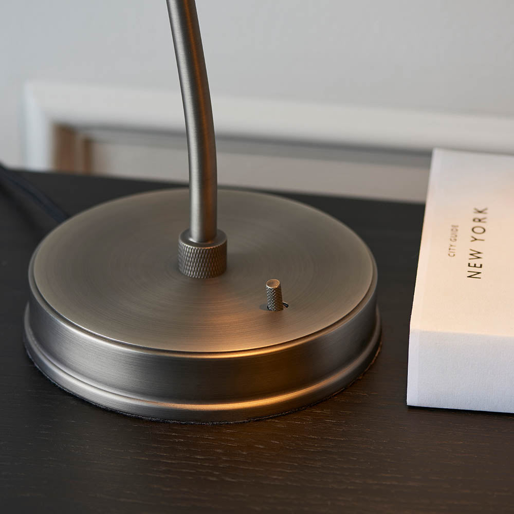 Close-up of the Endon Bramdale table lamp base in brushed silver with toggle switch and knurled knuckle, on a dark wood surface beside a book.