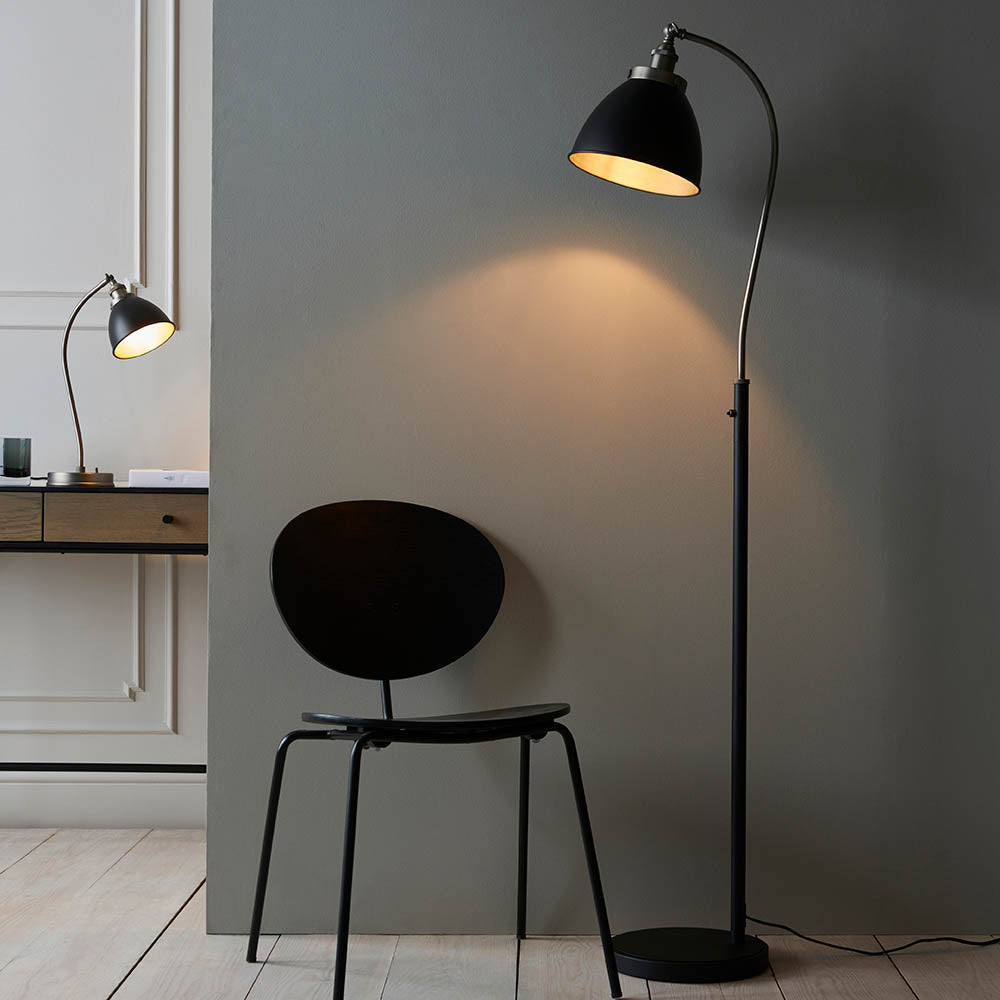 Bramdale Floor Lamp in silver and black, industrial-style adjustable metal shade on a curved arm, shown lit beside a black chair in a living room setting.
