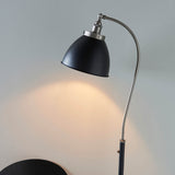 Bramdale Floor Lamp in silver and black, industrial-style adjustable arm with black metal shade, shown lit against a plain grey wall.