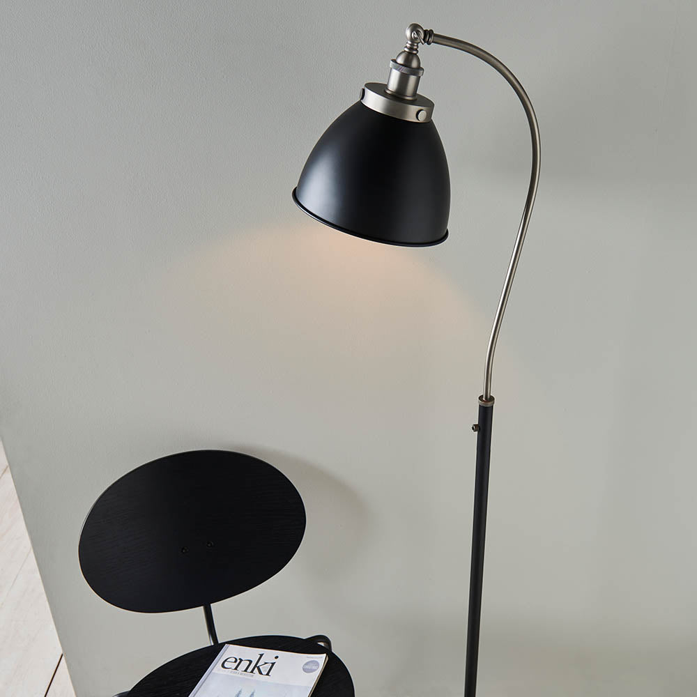 Bramdale Floor Lamp in silver and black with adjustable curved arm and rolled-edge metal shade, shown lit beside a chair in a living room setting.