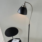 Bramdale Floor Lamp in silver and black with adjustable curved arm and rolled-edge metal shade, shown lit beside a chair in a living room setting.