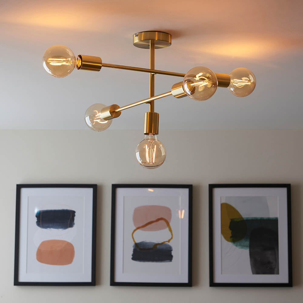 Endon Marlowe 5 light semi-flush ceiling light in satin brass with exposed E27 bulbs on angled arms, above three framed abstract prints on a wall.