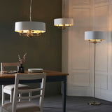 Endon Brenley V1 six-light pendants with white drum shades and brass fittings shown above a dining table beside a matching floor lamp in a panelled room.
