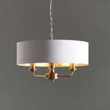 Endon Brenley 3 light pendant with a vintage white drum shade, gold metallic inner lining and antique brass fittings on a chain, against a grey background.