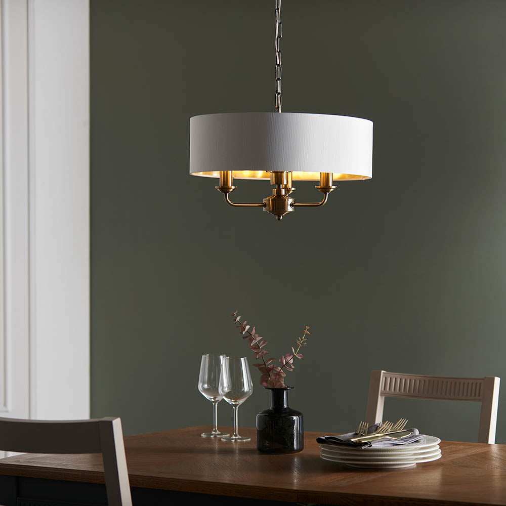 Endon Brenley 3 Light Pendant in antique brass with a white drum shade and gold-lined interior, hung above a wooden dining table against a dark green wall.