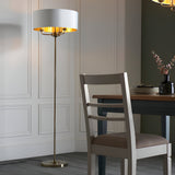 Endon Brenley 3 light floor lamp with white drum shade, gold inner lining and brass stem, beside a dining chair and table in a panelled room.