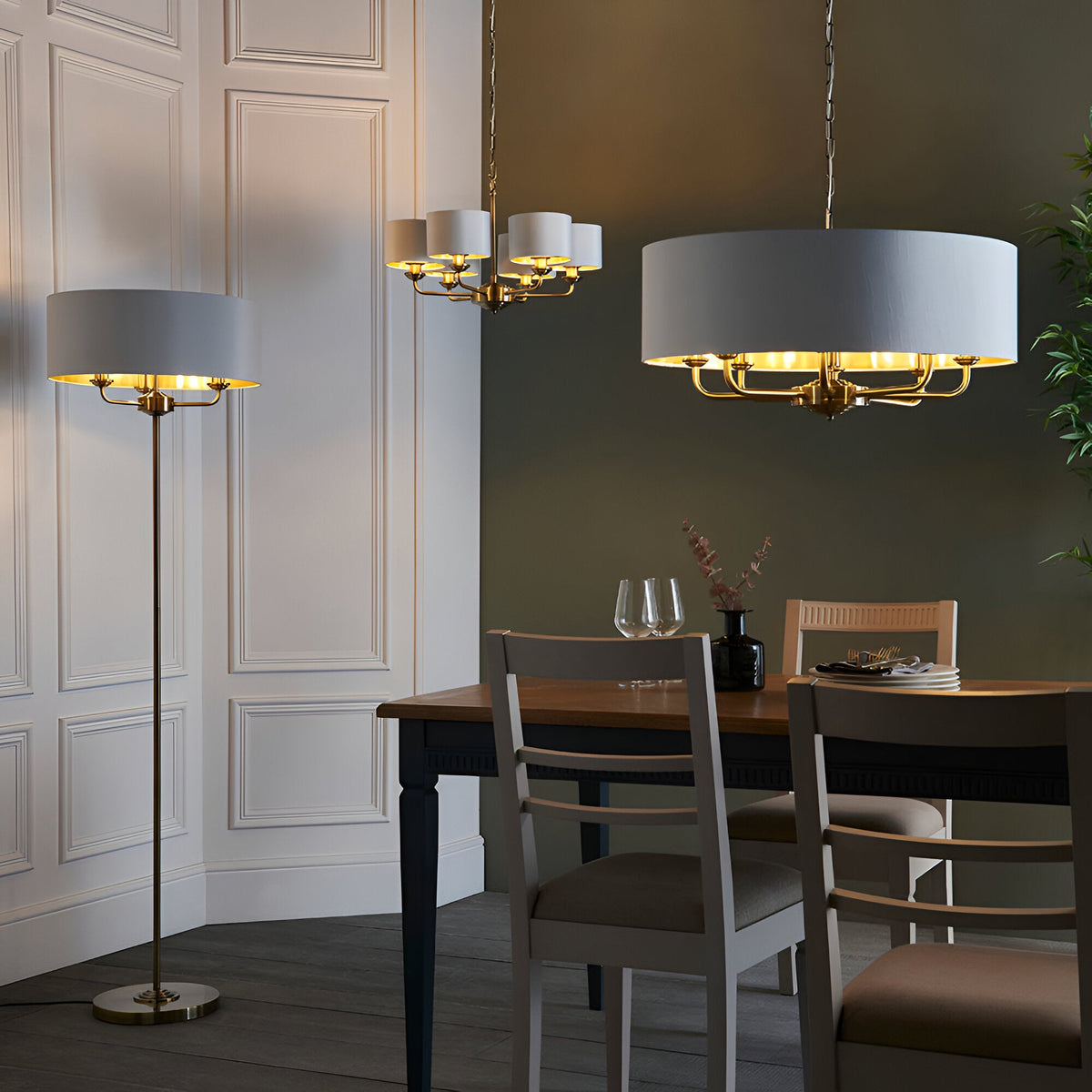 Endon Brenley floor lamp with wide white drum shade and brass metalwork, shown beside a dining table with matching pendant chandeliers in a panelled room.
