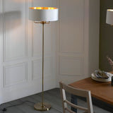 Endon Brenley 3 light floor lamp with brass stem and white drum shade lined in gold, shown beside a dining table against panelled walls.