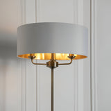 Endon Brenley 3 light floor lamp with white drum shade, gold metallic inner lining, and antique brass three-arm fitting against a panelled wall.