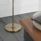 Close-up of the Endon Brenley floor lamp's round brass base and slim stem on grey wood flooring beside white panelled wainscoting.