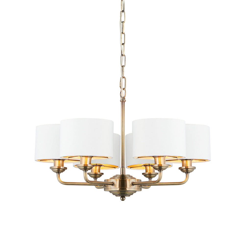 Brenley V2 six-light pendant with antique brass metalwork, six white fabric drum shades, and a chain suspension, shown on a plain white background.