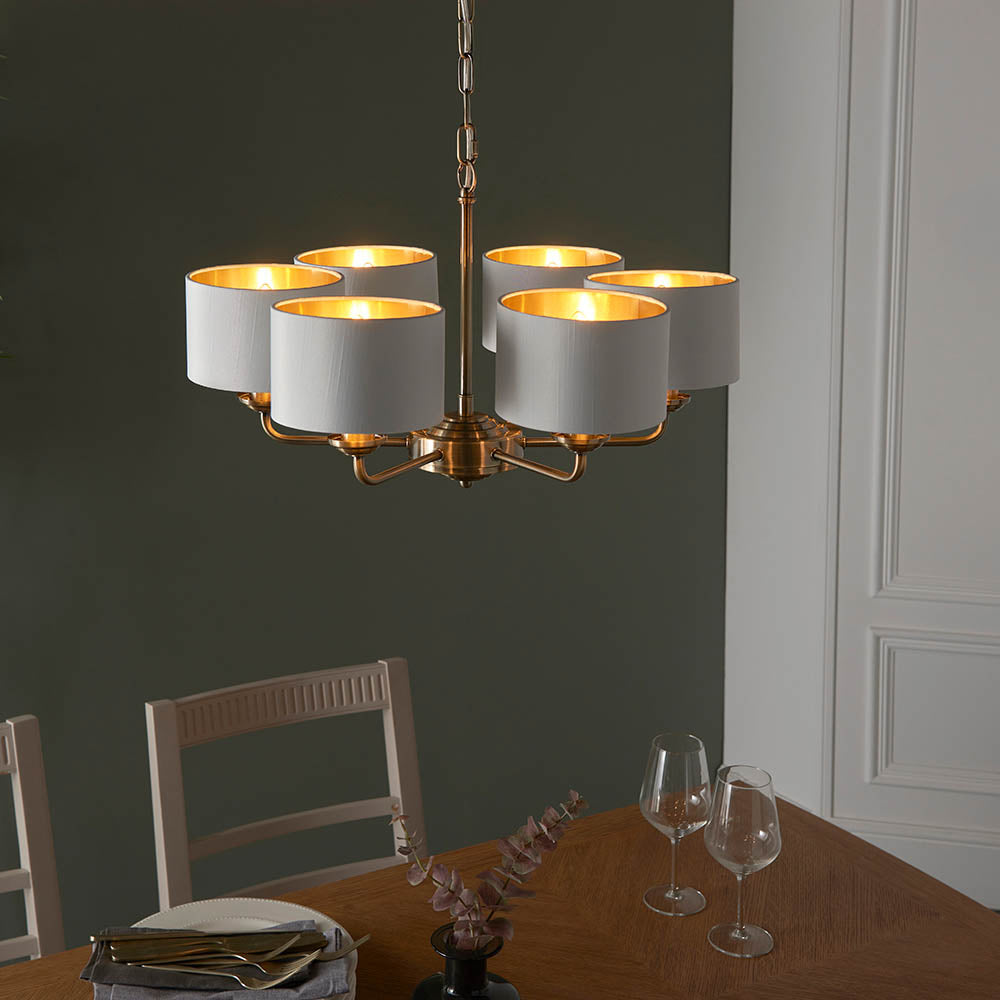 Brenley V2 six-light pendant with antique brass arms and white fabric shades with gold inner, shown above a dining table in a dark green room.