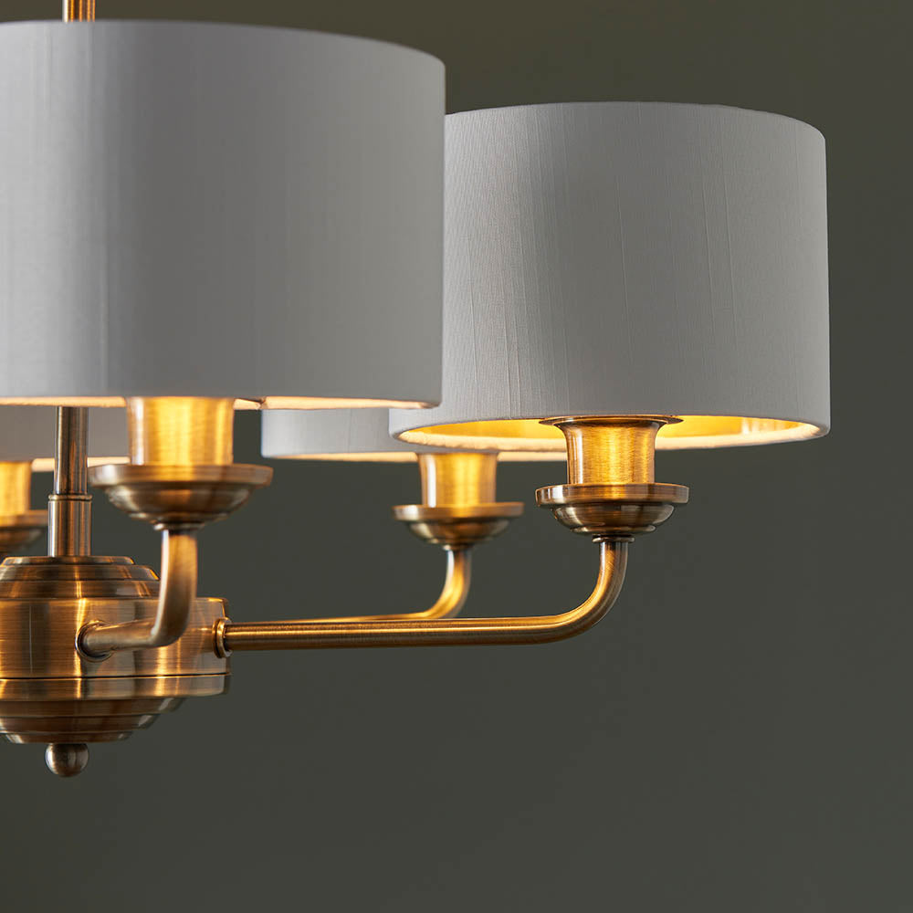 Brenley V2 6 Light Pendant close-up showing white fabric drum shades on brass arms with gold-lit interiors, set against a dark background.