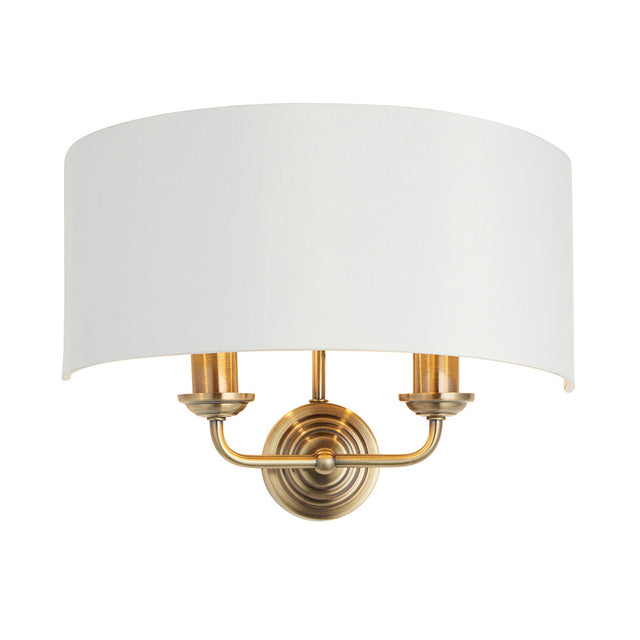 Endon Brenley 2 light wall light with a curved white linen half-shade on an antique brass twin-arm backplate, shown on a plain white background.
