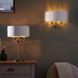 Endon Brenley 2 Light Wall Light in brass and gold with a white linen half-shade, illuminated above a table lamp on a sideboard in a dim living room.