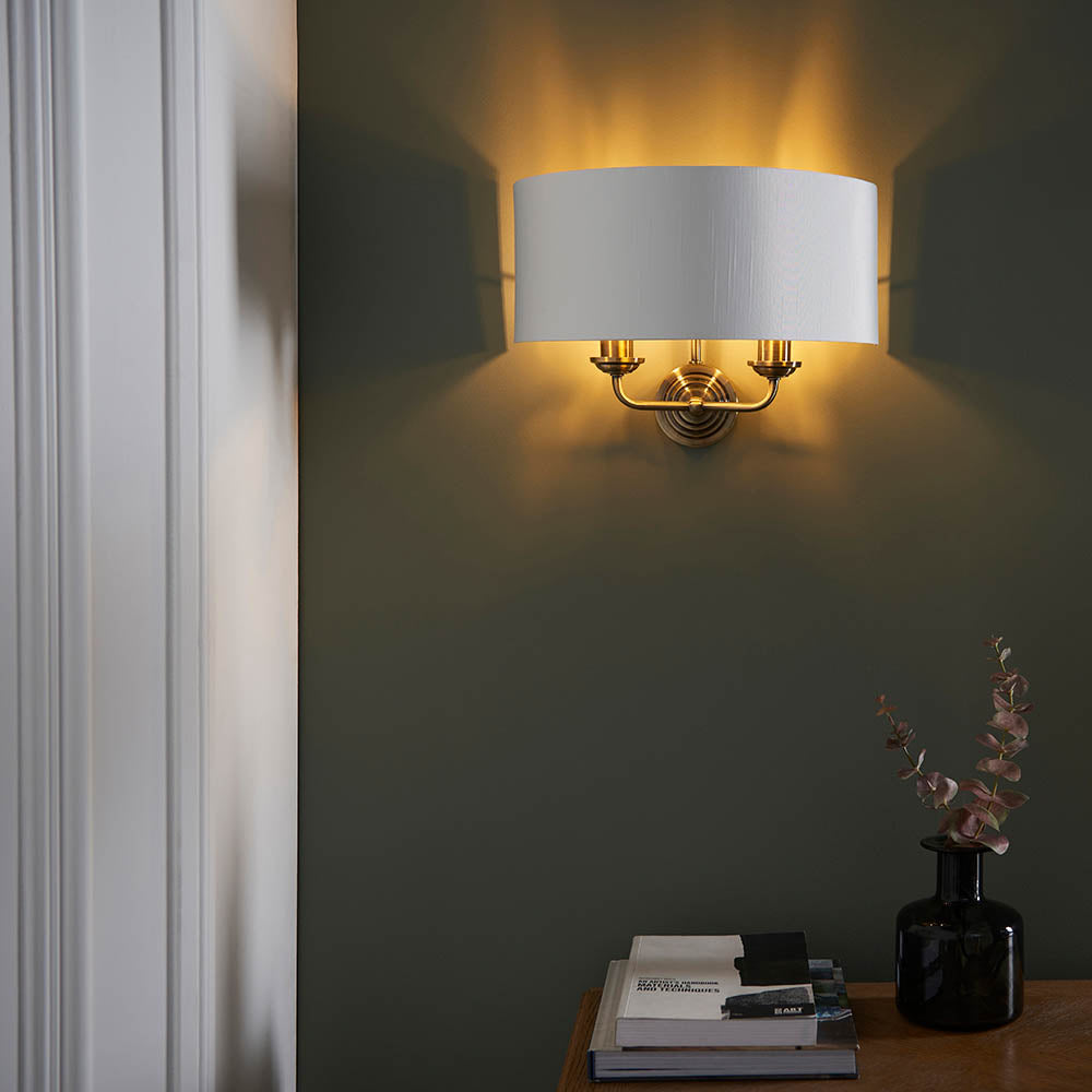 Endon Brenley 2 light wall light in brass and gold with a white linen half-shade, illuminated on a dark green wall above a sideboard with books and a vase.