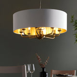 Endon Brenley 8 light pendant with white drum shade, gold-lined interior and brass arms, illuminated above a dining table against a dark wall.