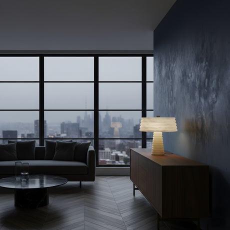 A modern living room with a dark sofa, round coffee table, and wooden sideboard topped with the Orivon 3 Light Sculpted Alabaster Table Lamp – Soft White. Large windows reveal a city skyline on a cloudy day.