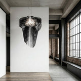 A modern, minimalist interior features the Aeris 5 Light Large Chandelier in black hanging from the ceiling, white walls, exposed brick, concrete floors, and tall industrial-style windows bringing in natural light.