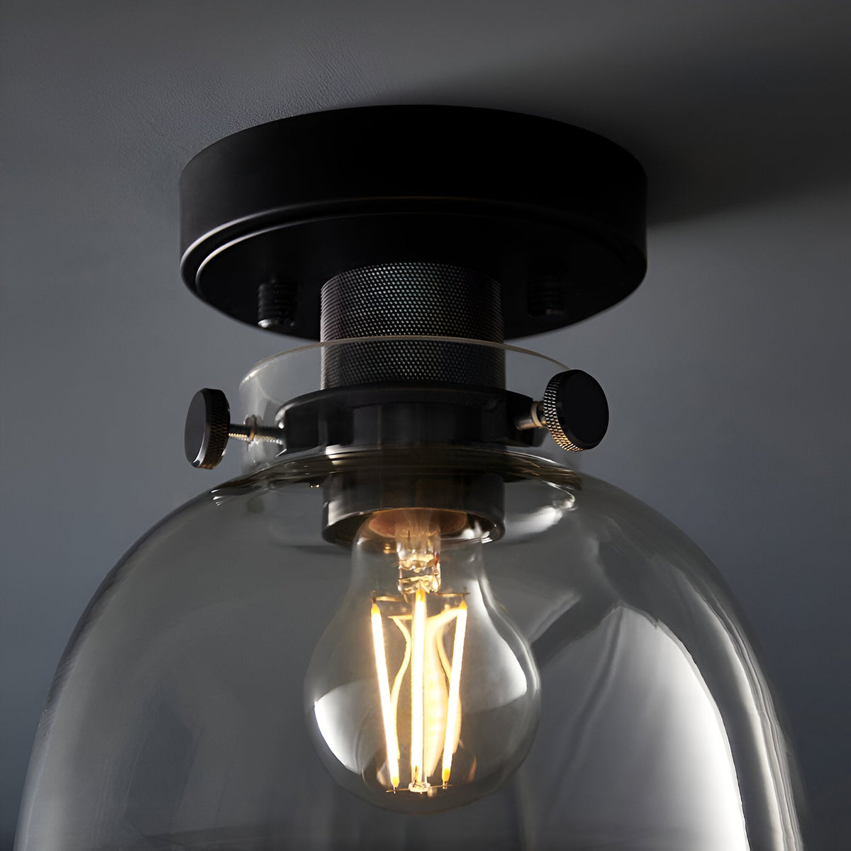 Endon Camden matt black flush ceiling light shown close-up with knurled thumbscrews securing a clear glass shade around a lit E27 filament bulb.