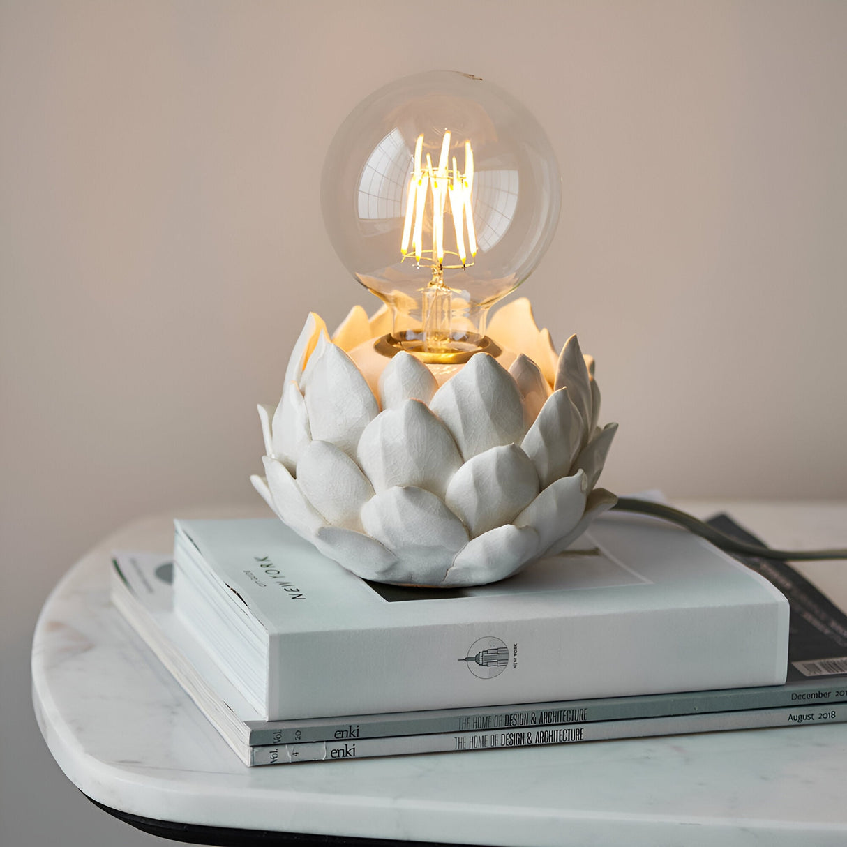 Aldgrove Table Lamp in Antique Brass with an oatmeal crackle-glazed ceramic artichoke base, lit E27 globe bulb, on stacked books atop a marble table.