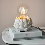 Aldgrove Table Lamp in Antique Brass with an oatmeal crackle-glazed ceramic artichoke base, lit E27 globe bulb, on stacked books atop a marble table.