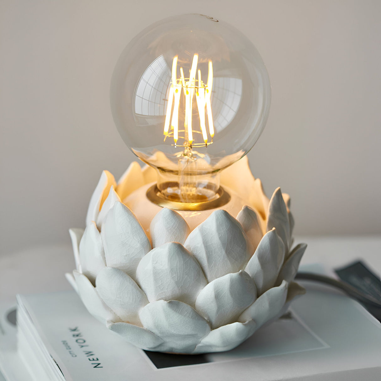 Aldgrove Table Lamp with white ceramic artichoke-style layered petal base, antique brass neck detail, and lit E27 globe bulb, shown on a book.