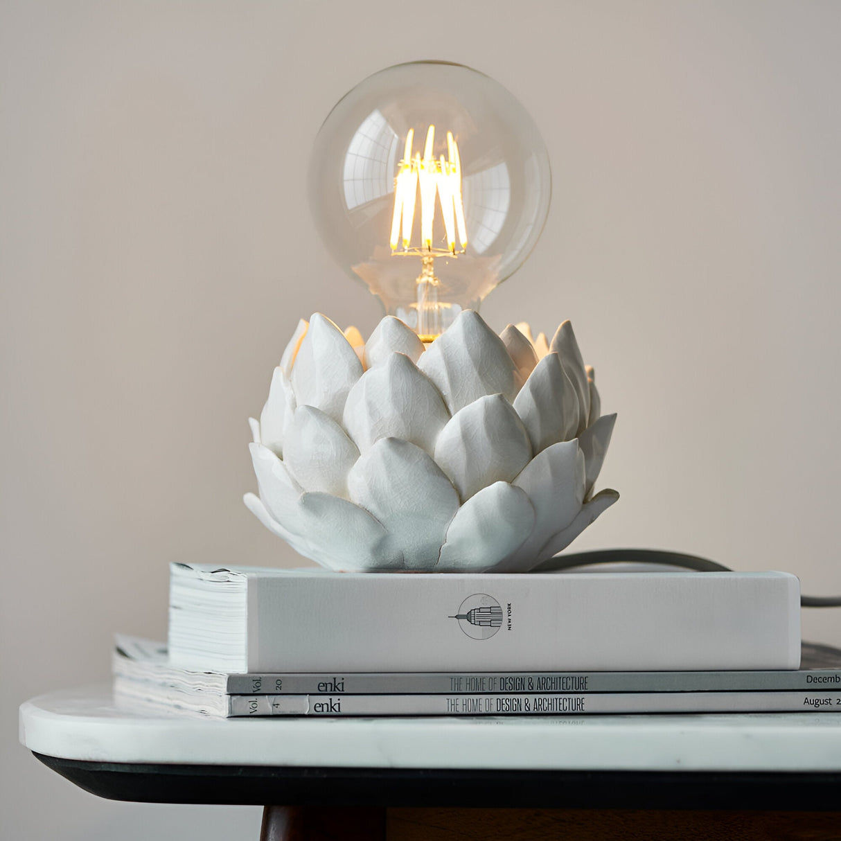 Aldgrove Table Lamp in white ceramic artichoke design with antique brass neck detail, shown lit with a clear E27 bulb atop stacked books.