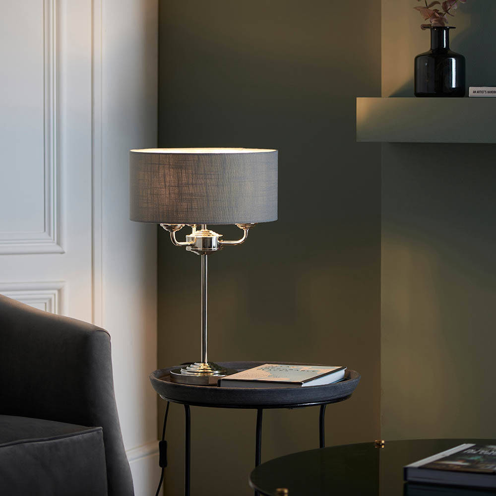 Endon Brenley 3 Light Table Lamp in nickel with a grey linen drum shade, styled on a dark round side table in a green-toned living room.