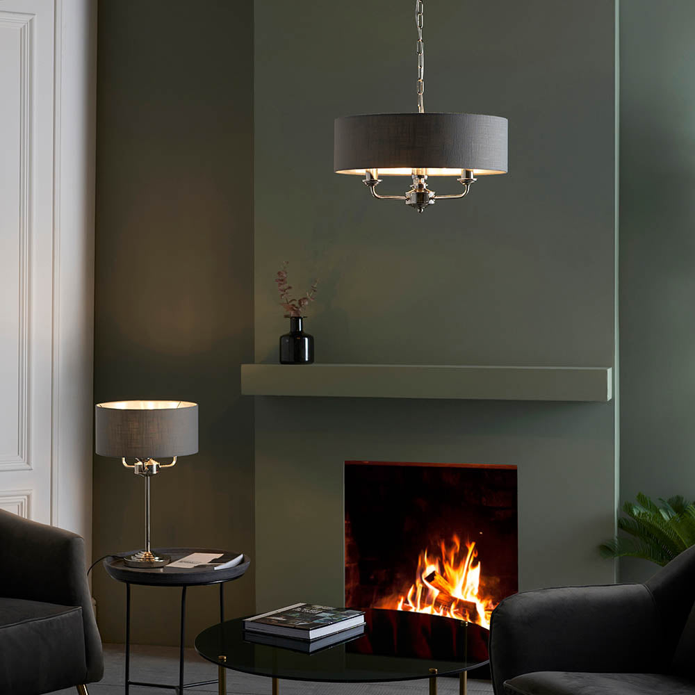 Endon Brenley 3 Light Table Lamp in nickel with a grey linen shade, lit on a side table beside a fireplace with a matching pendant above the mantel.