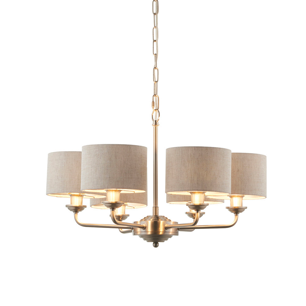 Brenley 6-light pendant with brass fittings and six curved arms holding natural linen drum shades, suspended from a chain, on a plain white background.