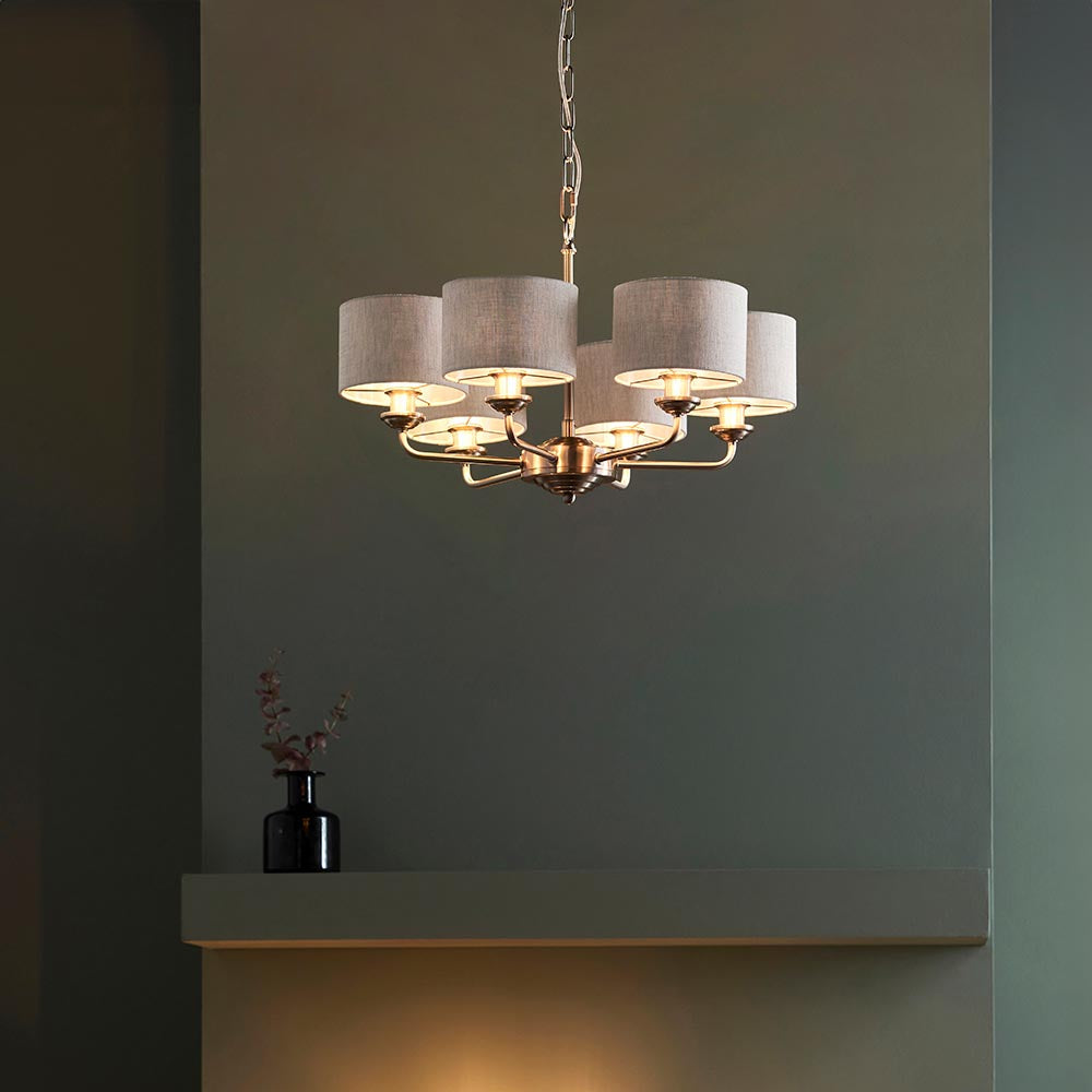 Brenley 6-light pendant with brass arms and grey fabric drum shades, hung from a chain in a room with a dark green wall and shelf.