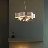 Brenley 6-light pendant with brass arms and grey fabric drum shades, hung from a chain in a room with a dark green wall and shelf.