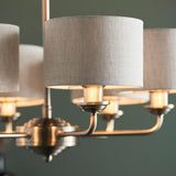 Close-up of the Brenley 6 Light Pendant in brass and chrome, showing multiple arms with grey linen drum shades and lit E14 bulbs against a green wall.