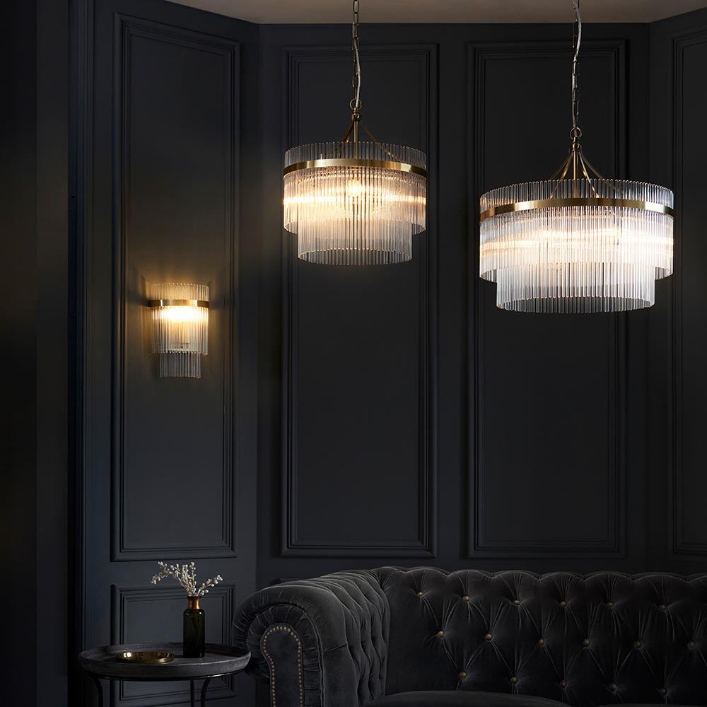 Endon Beaumont 5 Light Pendant in brass with tiered clear glass rods, shown alongside a matching wall light above a dark tufted sofa in a panelled room.